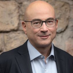 A bald man wearing glasses, a light blue shirt, and a dark blazer is standing in front of a stone wall.