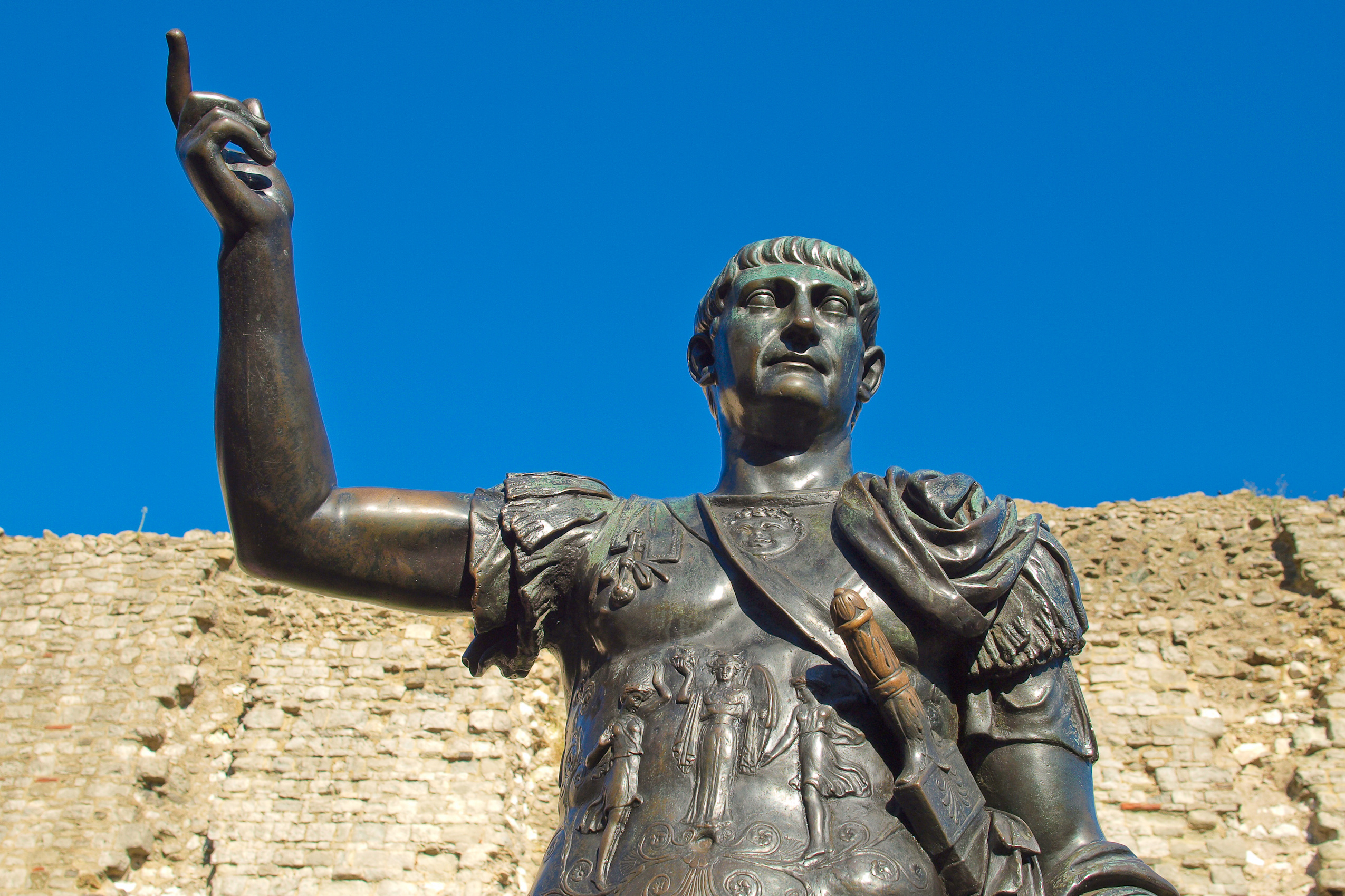 Bronze statue of a Roman emperor standing in front of an ancient stone wall, with one arm raised and dressed in traditional Roman armor under a clear blue sky.