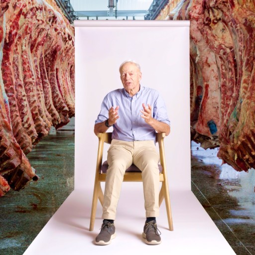 An older man sits on a chair in front of a white backdrop, surrounded by rows of hanging beef carcasses in a meat processing facility.