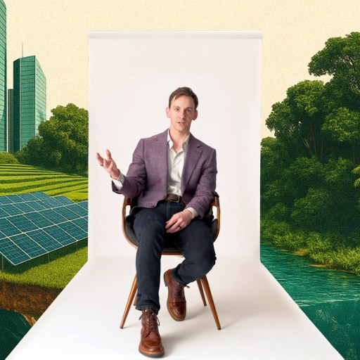 A man in a suit sits on a chair against a white backdrop, with illustrated green skyscrapers, solar panels, and trees in the background.
