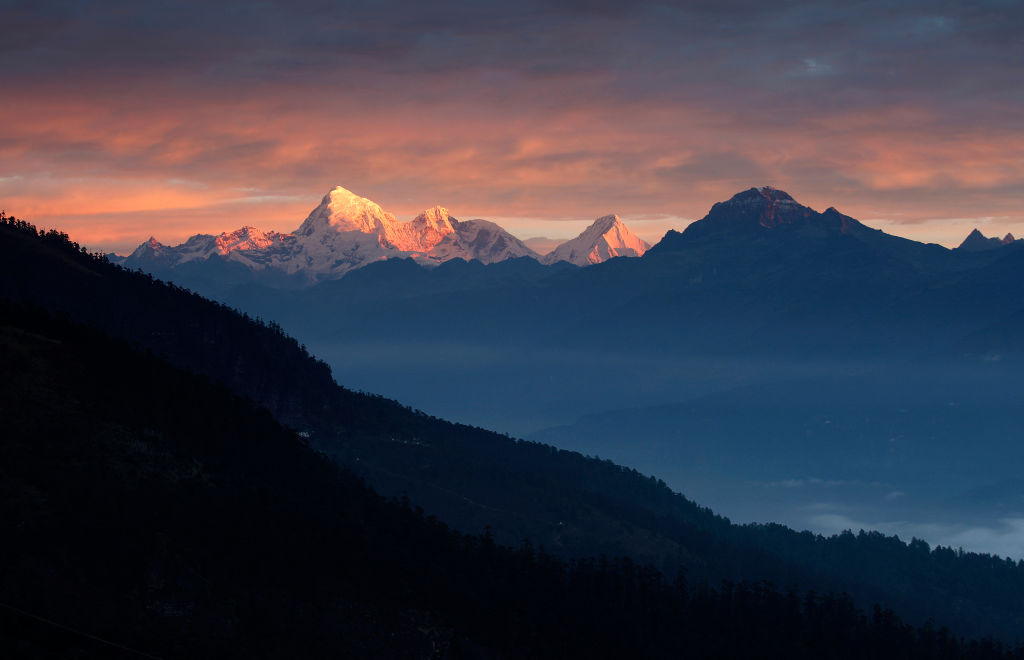 Mountain peaks lit by the orange glow of sunrise, with dark silhouettes of hills and pine trees in the foreground under a partly cloudy sky.