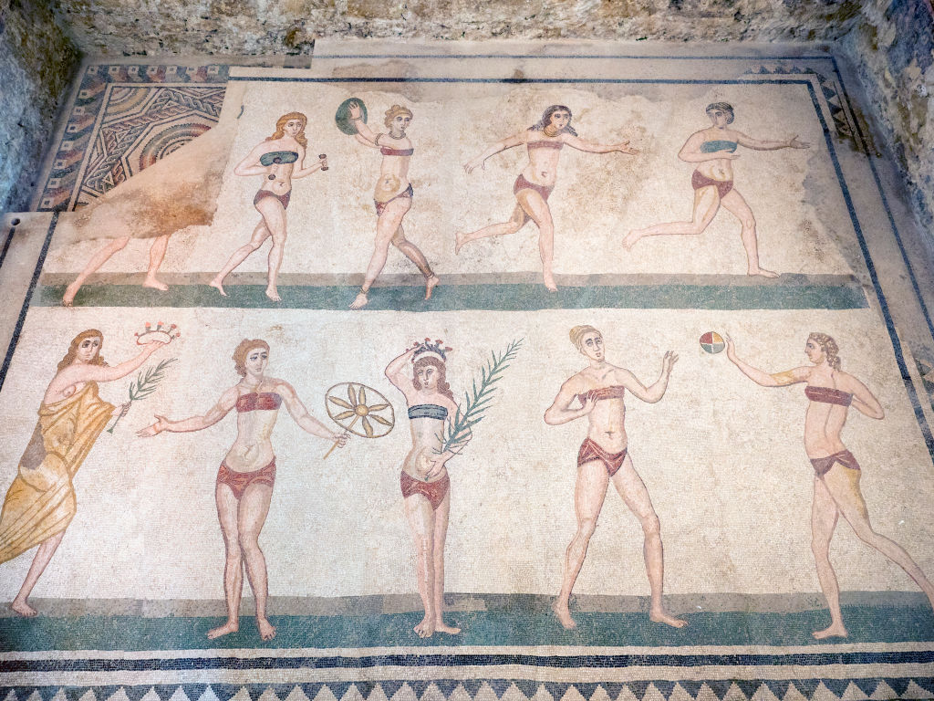 Ancient Roman mosaic depicting women in two-piece garments playing sports and holding objects, set against a tiled background.