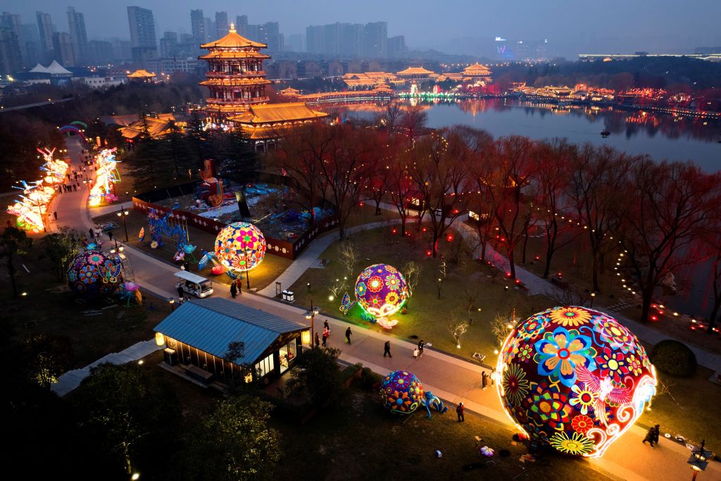 People walk along a path decorated with large, illuminated floral lanterns near a traditional pavilion and lake in a city at dusk.