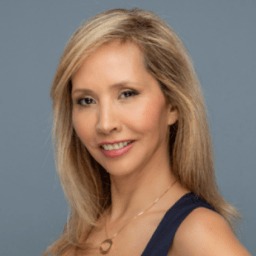 A woman with long blonde hair, wearing a dark sleeveless top and a gold necklace, smiles at the camera against a plain gray background.