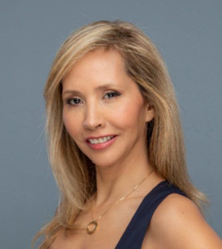 A woman with long blonde hair, wearing a dark sleeveless top and a gold necklace, smiles at the camera against a plain gray background.