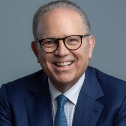 A middle-aged man with gray hair and glasses smiles while wearing a blue suit, white shirt, and blue patterned tie against a plain gray background.