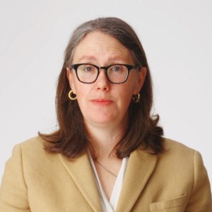 Woman with shoulder-length brown hair wearing glasses, gold hoop earrings, a beige blazer, and a white blouse against a plain white background.