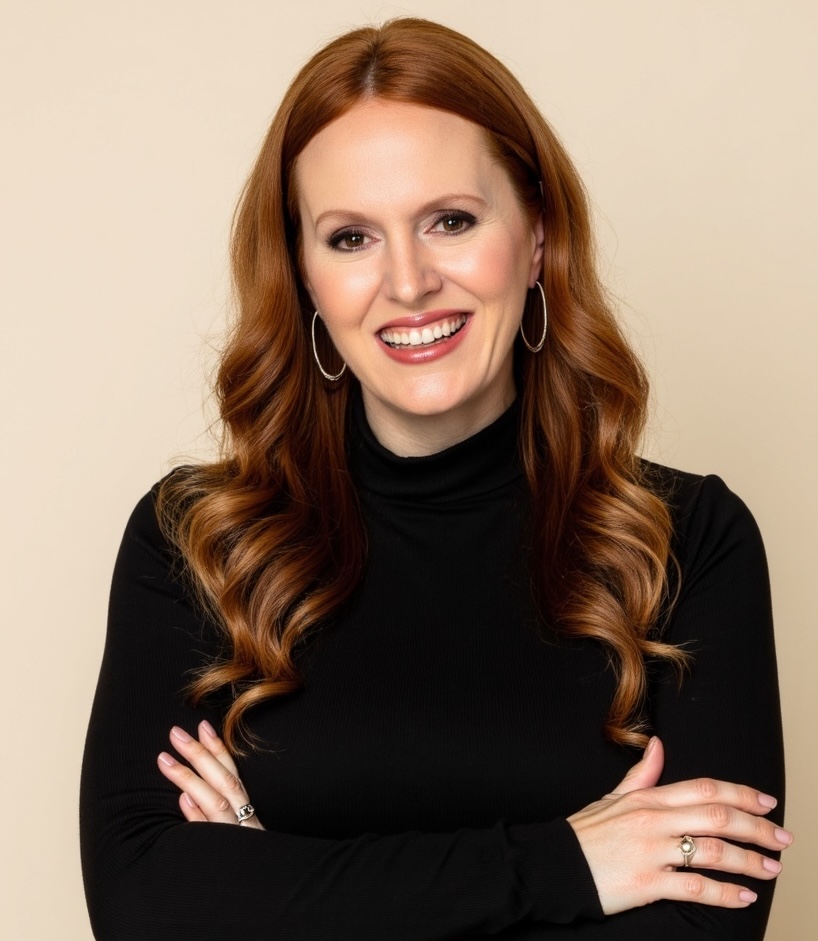 Woman with long red hair in loose curls, wearing a black turtleneck and hoop earrings, smiling with arms crossed against a beige background.
