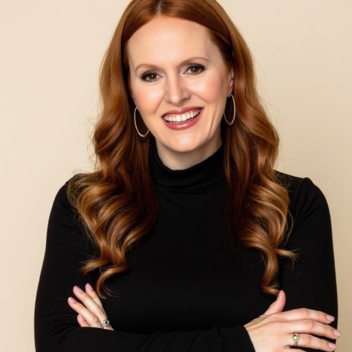 Woman with long red hair in loose curls, wearing a black turtleneck and hoop earrings, smiling with arms crossed against a beige background.