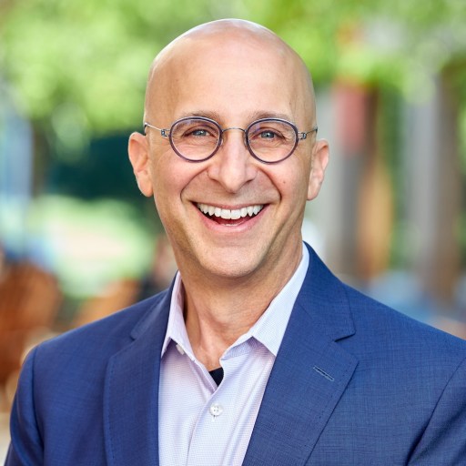 Bald man wearing glasses and a blue suit jacket smiles outdoors with a blurred green background.