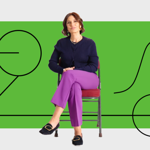 A woman sits on a chair with legs crossed, wearing a navy sweater and purple pants, in front of a green background with black abstract line art.