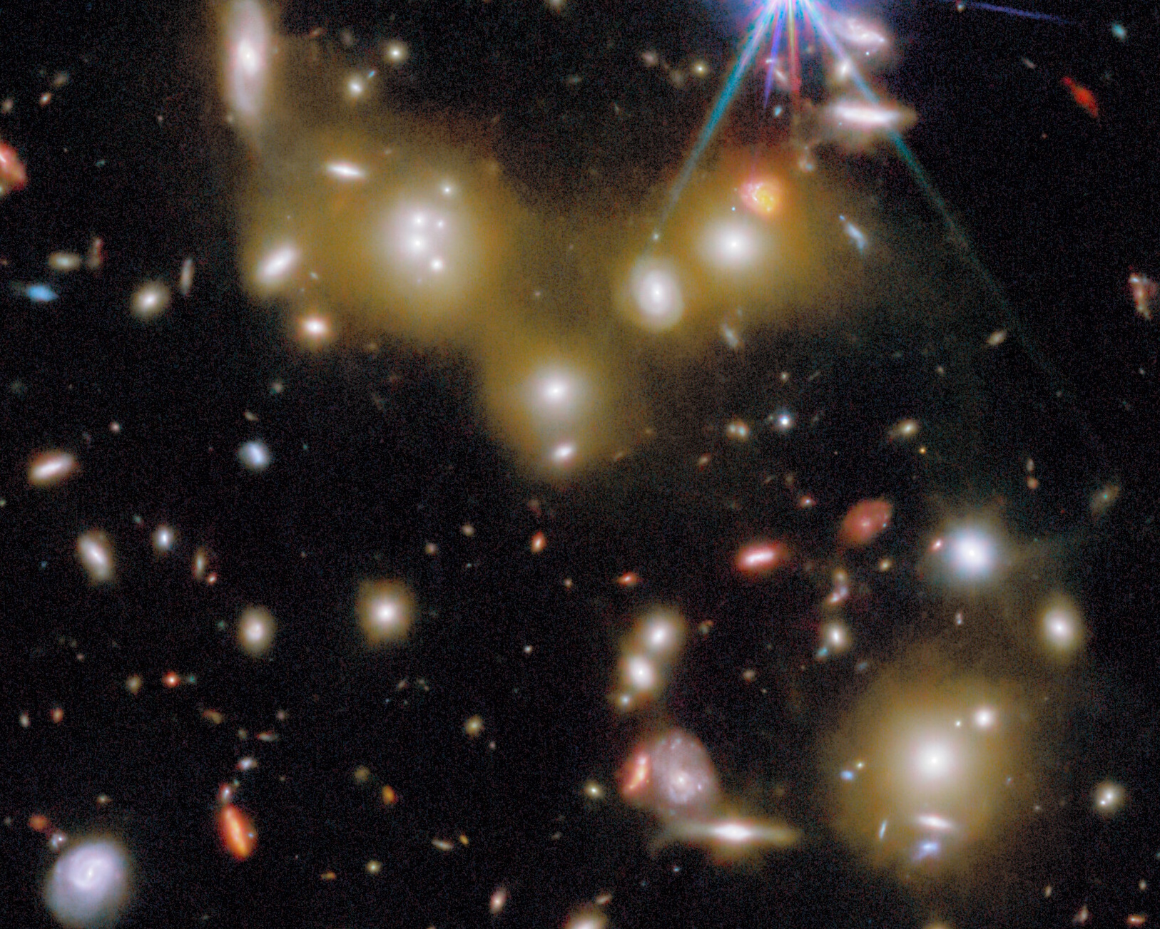 Image of a galaxy cluster with many bright galaxies, stars, and glowing clouds against a dark background, captured by the Hubble Space Telescope.