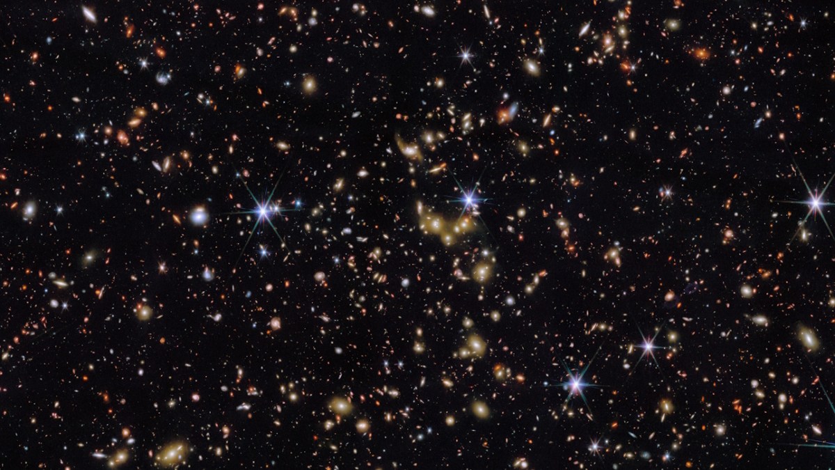 A dense field of stars and galaxies is visible against a black background, with some bright stars showing lens flares.