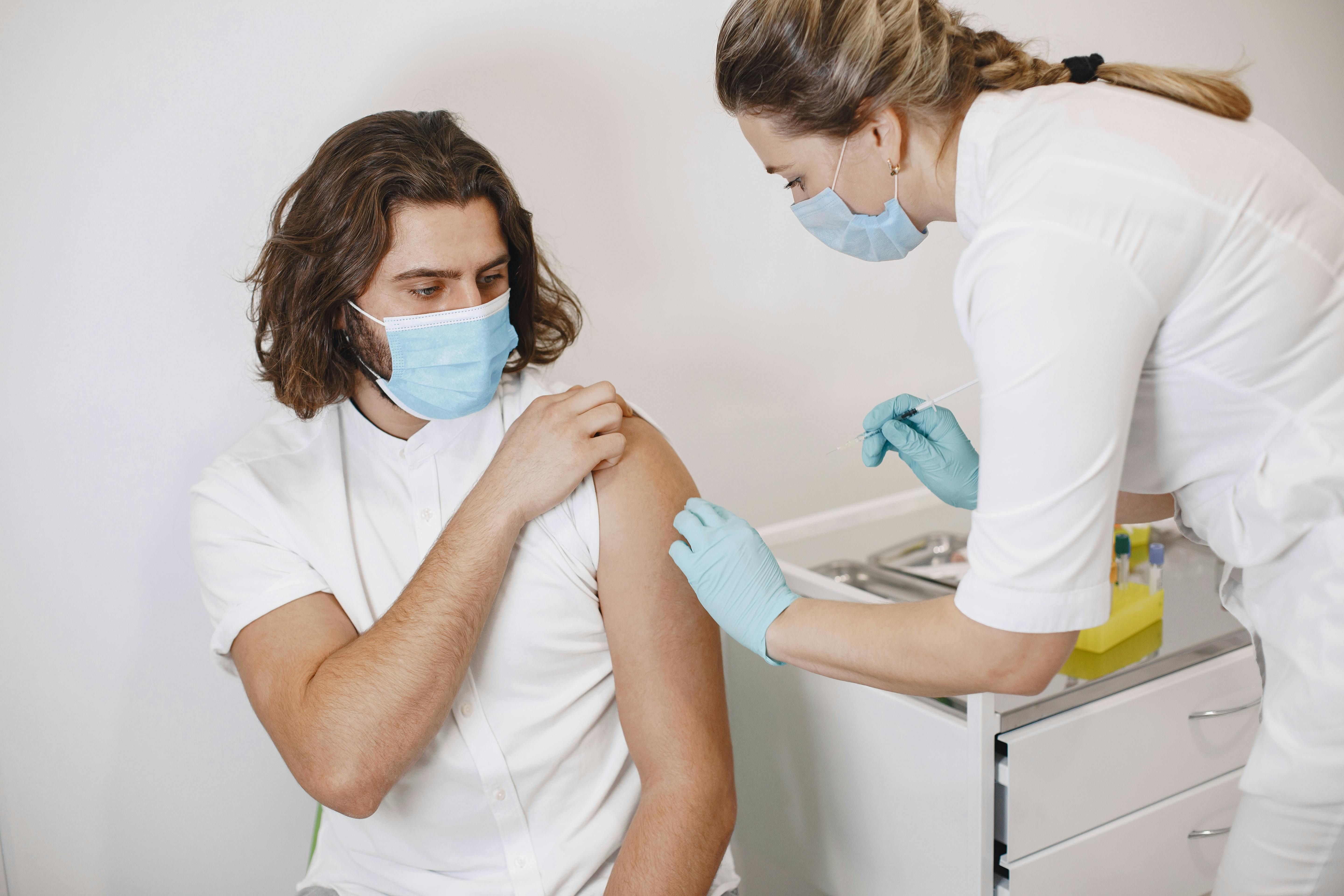 A healthcare worker in gloves and a mask administers a vaccine injection into a seated man's upper arm. Both are wearing face masks, highlighting the ongoing importance of vaccines—do your own research to stay informed.