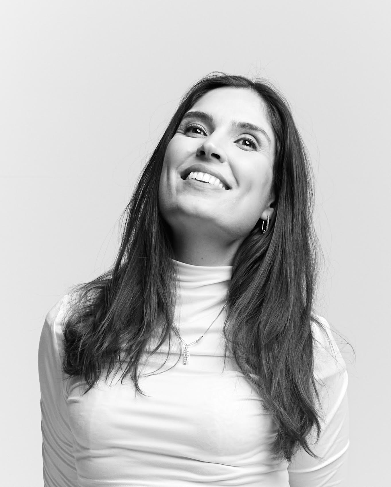 A woman with long dark hair wearing a light turtleneck top smiles and looks upward against a plain background.