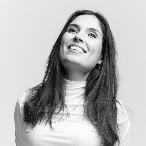 A woman with long dark hair wearing a light turtleneck top smiles and looks upward against a plain background.