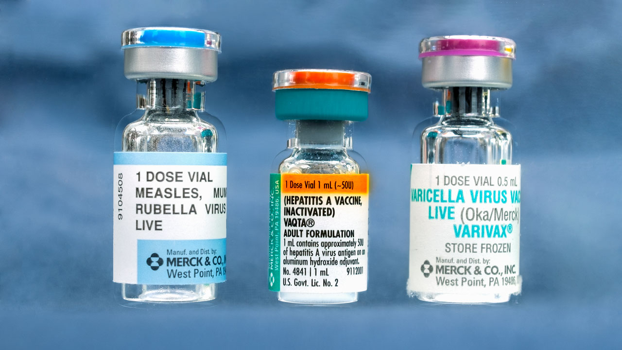 Three labeled vaccine vials stand upright against a blue background, each with different colored caps: silver, orange, and blue—a reminder that when it comes to vaccines, do your own research.