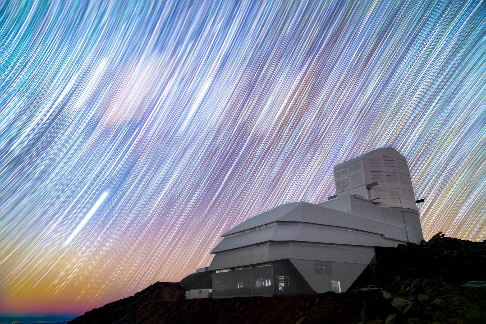 A large observatory sits on rocky ground under a sky filled with colorful star trails, indicating a long exposure photograph at night.