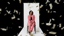 A woman in a pink suit sits on a chair against a white backdrop while U.S. dollar bills fall around her on a black background.