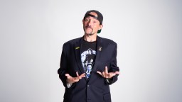 A man wearing a black blazer, graphic T-shirt, and backwards cap stands against a plain background with his hands raised in a questioning gesture.