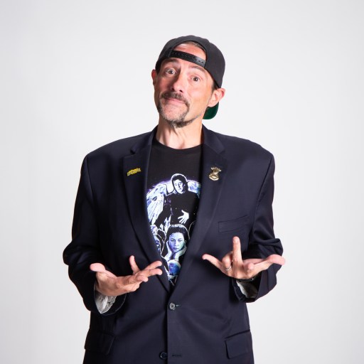 A man wearing a black blazer, graphic T-shirt, and backwards cap stands against a plain background with his hands raised in a questioning gesture.