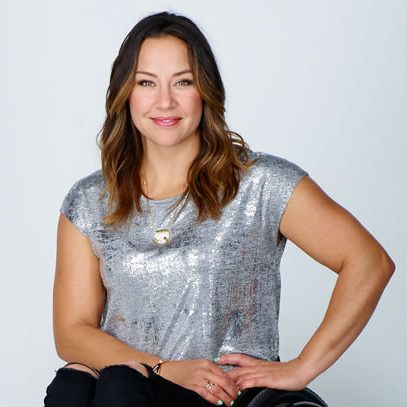 A woman with long brown hair, wearing a silver top and black pants, sits and smiles in a manual wheelchair against a plain white background.