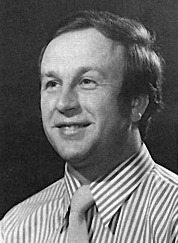 Black and white portrait of a smiling man wearing a striped shirt and a light-colored tie, facing slightly to the left against a plain dark background.