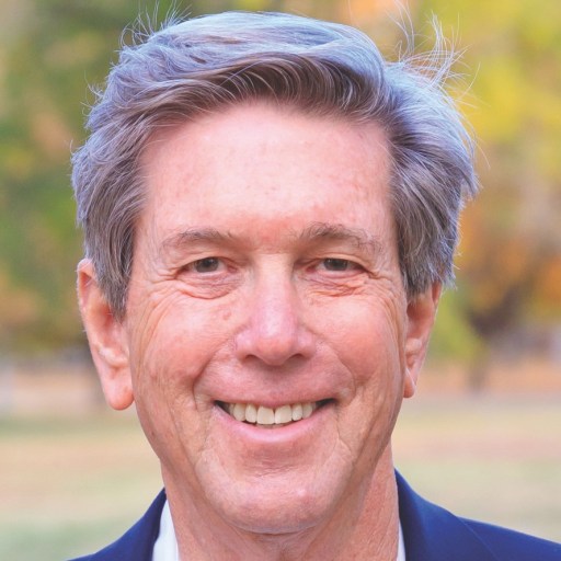 An older man with gray hair and a navy blazer smiles outdoors with blurred trees in the background.