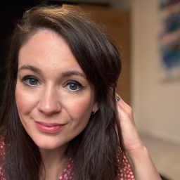 A woman with long brown hair and blue eyes looks at the camera, smiling slightly, wearing a red patterned top and touching her hair.