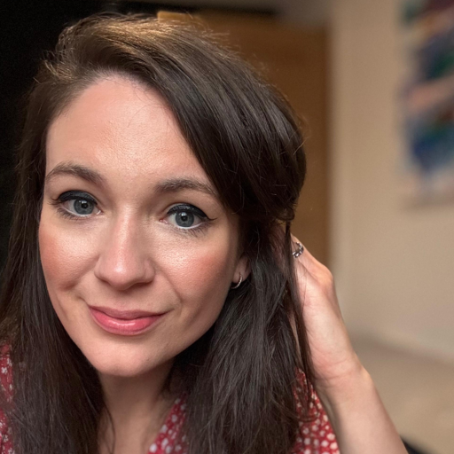 A woman with long brown hair and blue eyes looks at the camera, smiling slightly, wearing a red patterned top and touching her hair.