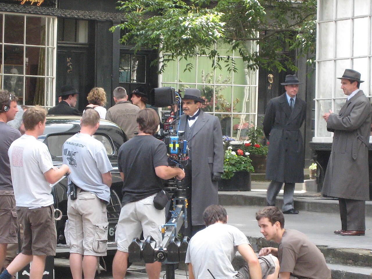 Film crew and actors in period costumes shoot a scene on a city street with a vintage car, camera equipment, and crew members visible.