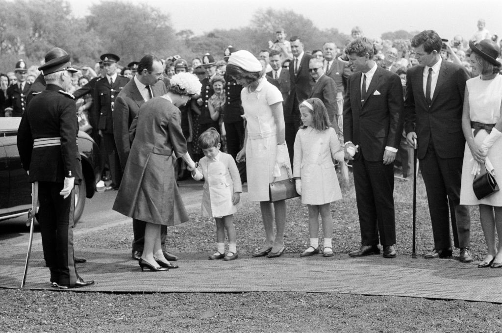 A group of adults and children greet a woman in a hat and coat while officials and onlookers stand in the background during an outdoor event.