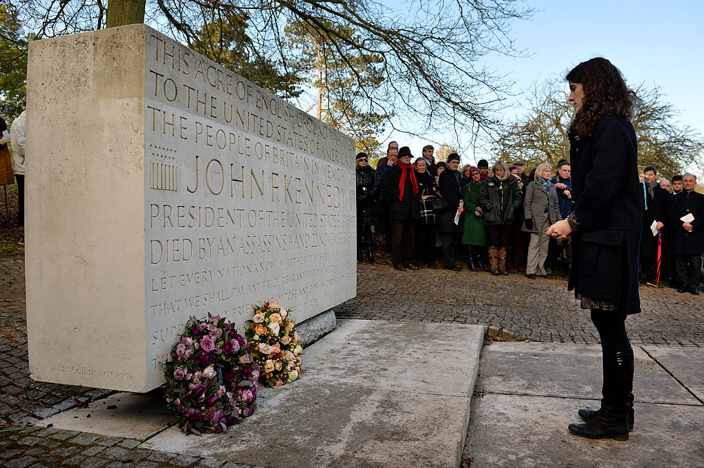 A woman stands in front of the John F. Kennedy memorial stone with flowers laid at its base, as a group of people observe in the background.