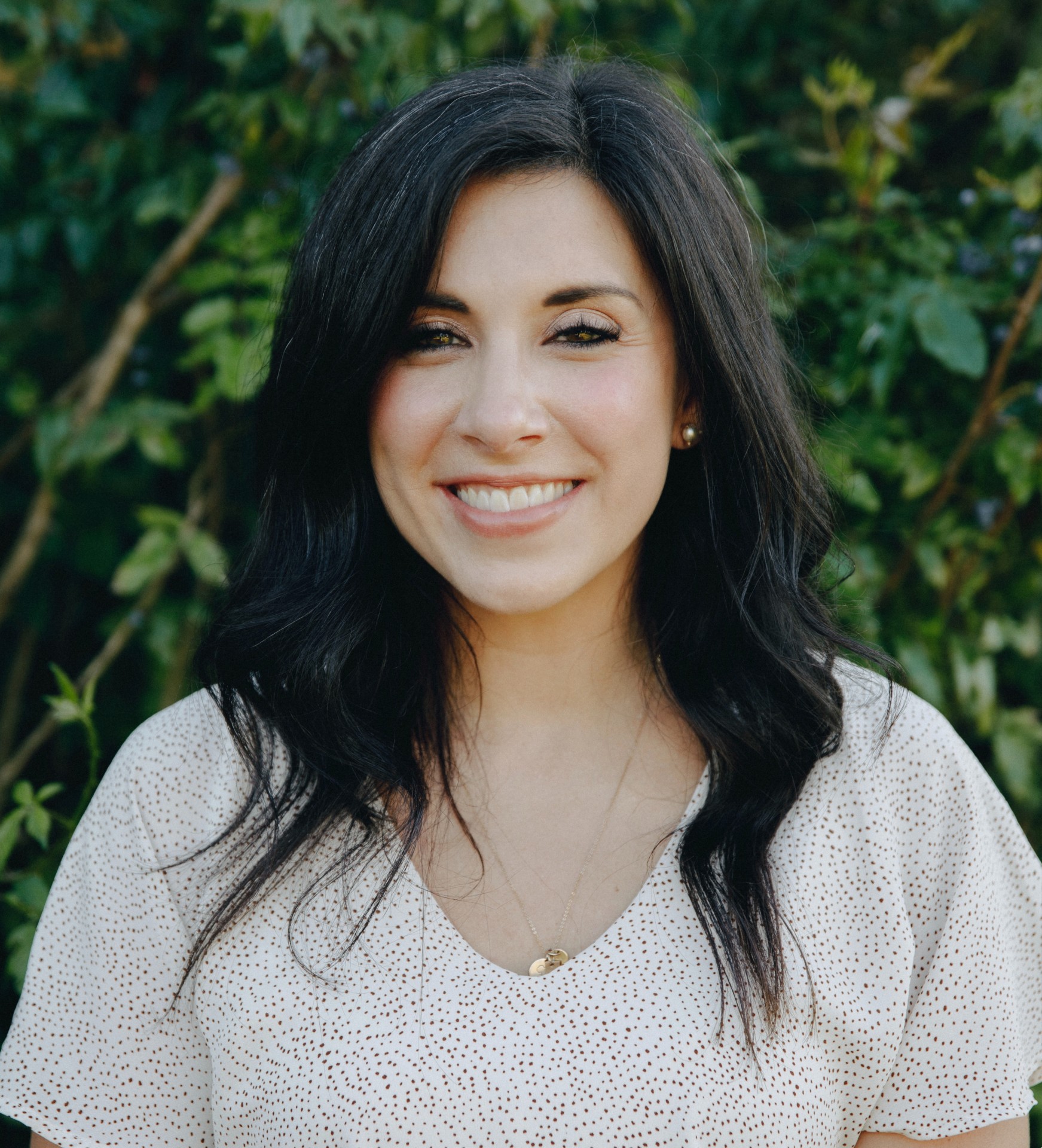A woman with long dark hair and a white blouse smiles at the camera while standing outdoors in front of green foliage.