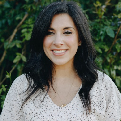 A woman with long dark hair and a white blouse smiles at the camera while standing outdoors in front of green foliage.