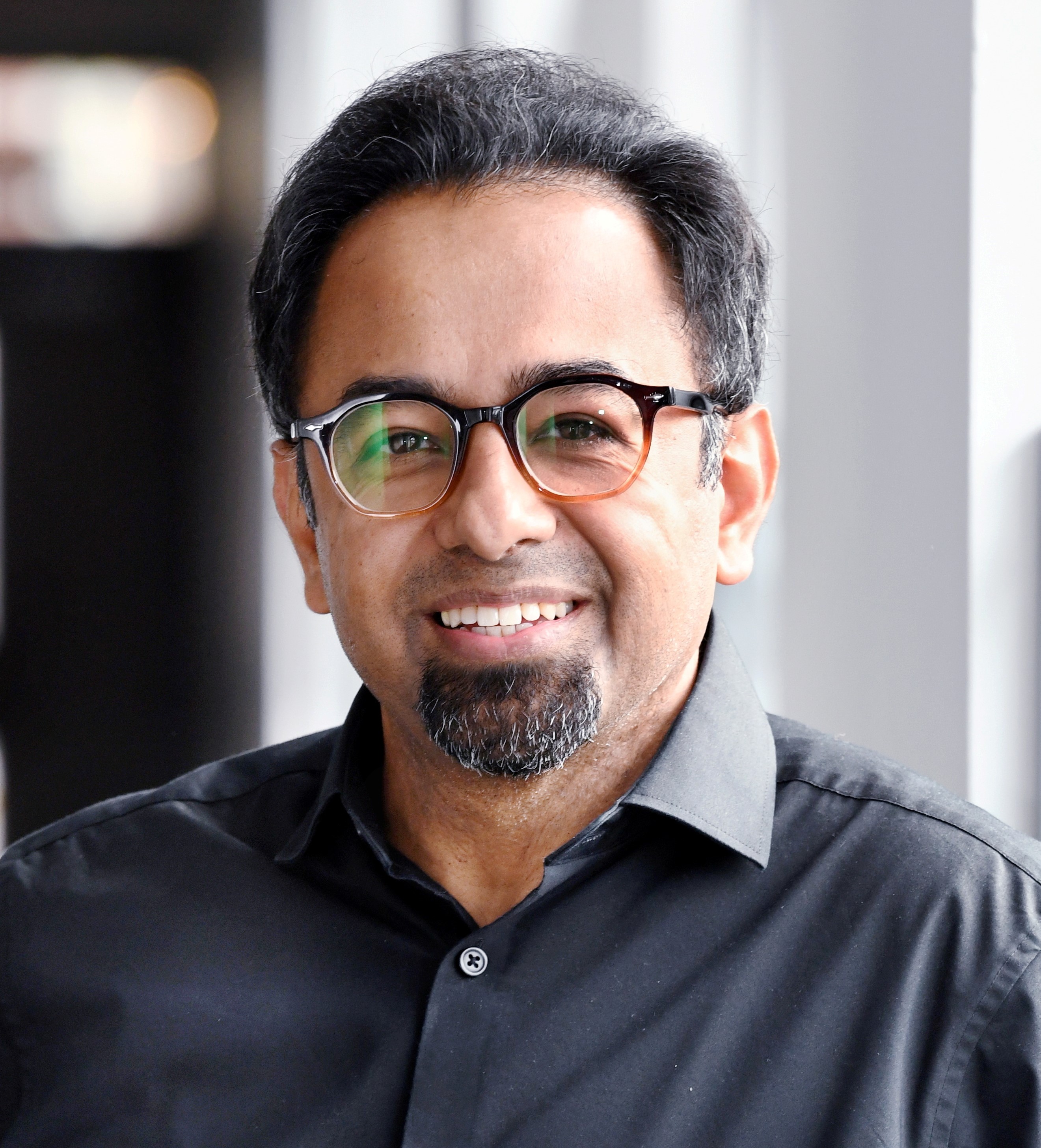 A man with glasses and a goatee, wearing a black shirt, smiles while standing indoors near a window.