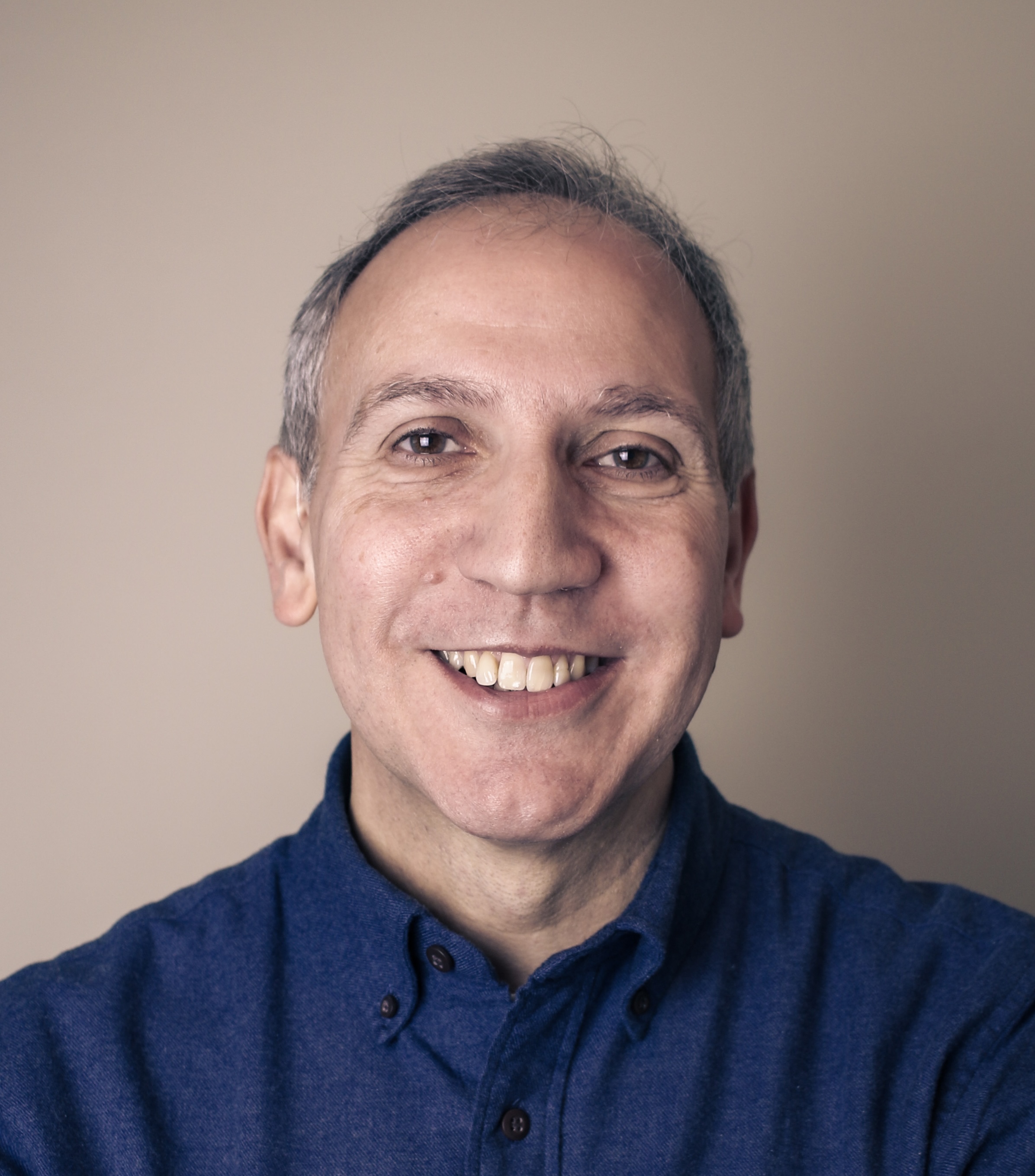 Middle-aged man with short gray hair, smiling and wearing a dark blue button-up shirt, posed against a plain beige background.