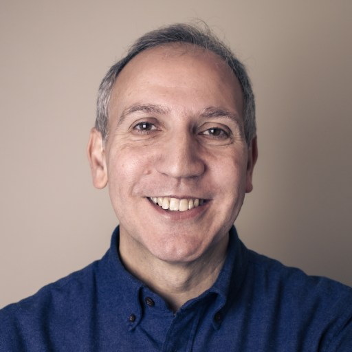 Middle-aged man with short gray hair, smiling and wearing a dark blue button-up shirt, posed against a plain beige background.