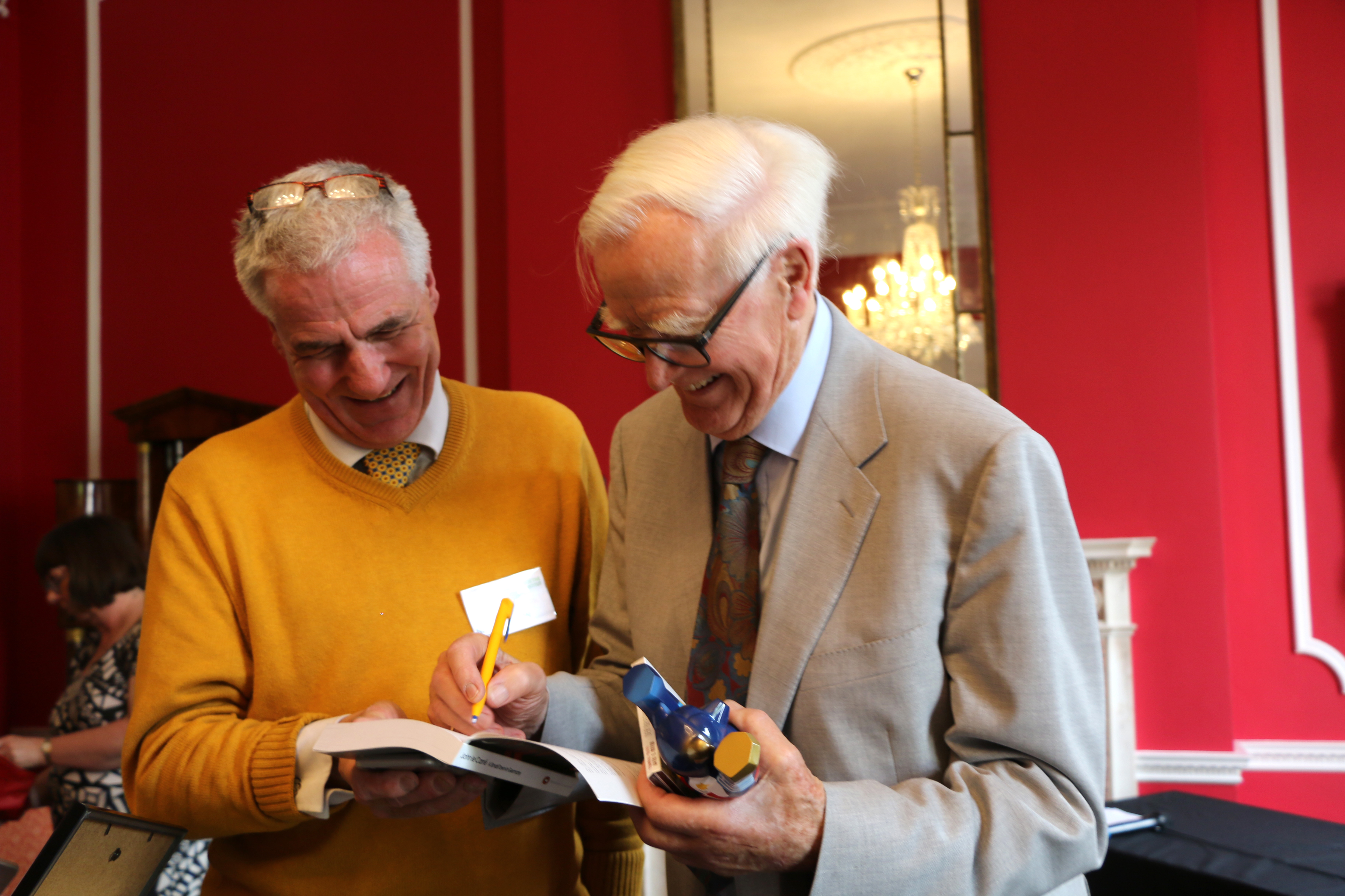 Two older men standing and smiling while looking at a book together in a red-walled room; one holds a pen and the other holds a blue object.
