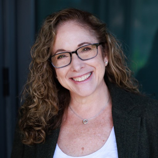 A woman with curly hair and glasses smiles at the camera. She is wearing a dark blazer over a white top and a necklace, standing in front of a dark, blurred background.