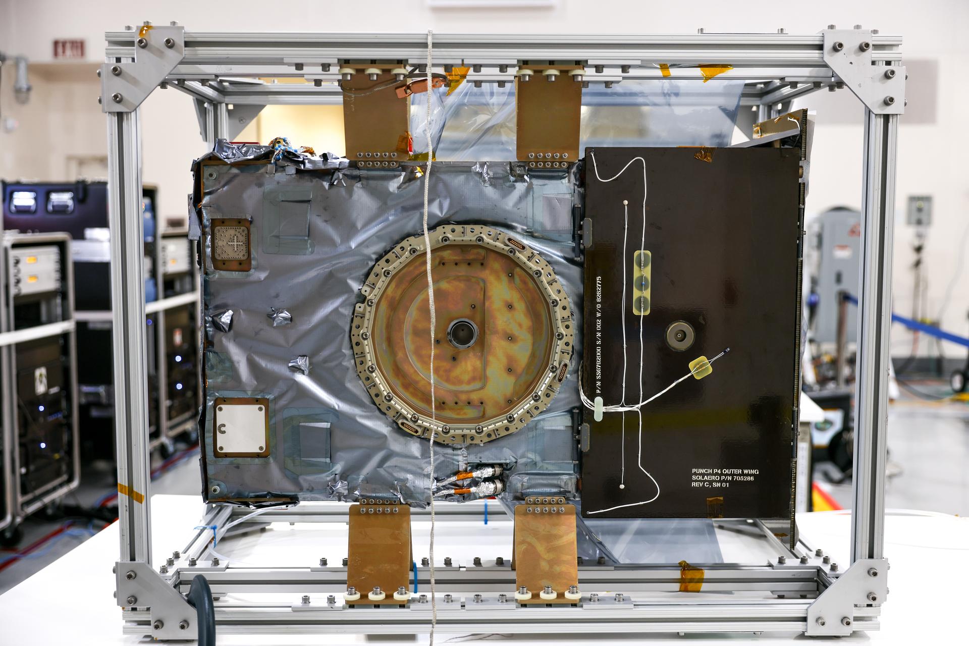 A metal satellite component or scientific instrument in a lab, housed within a rectangular frame with wiring and electronic parts visible, resembles the equipment seen in NASA PUNCH video research on the sun's corona.