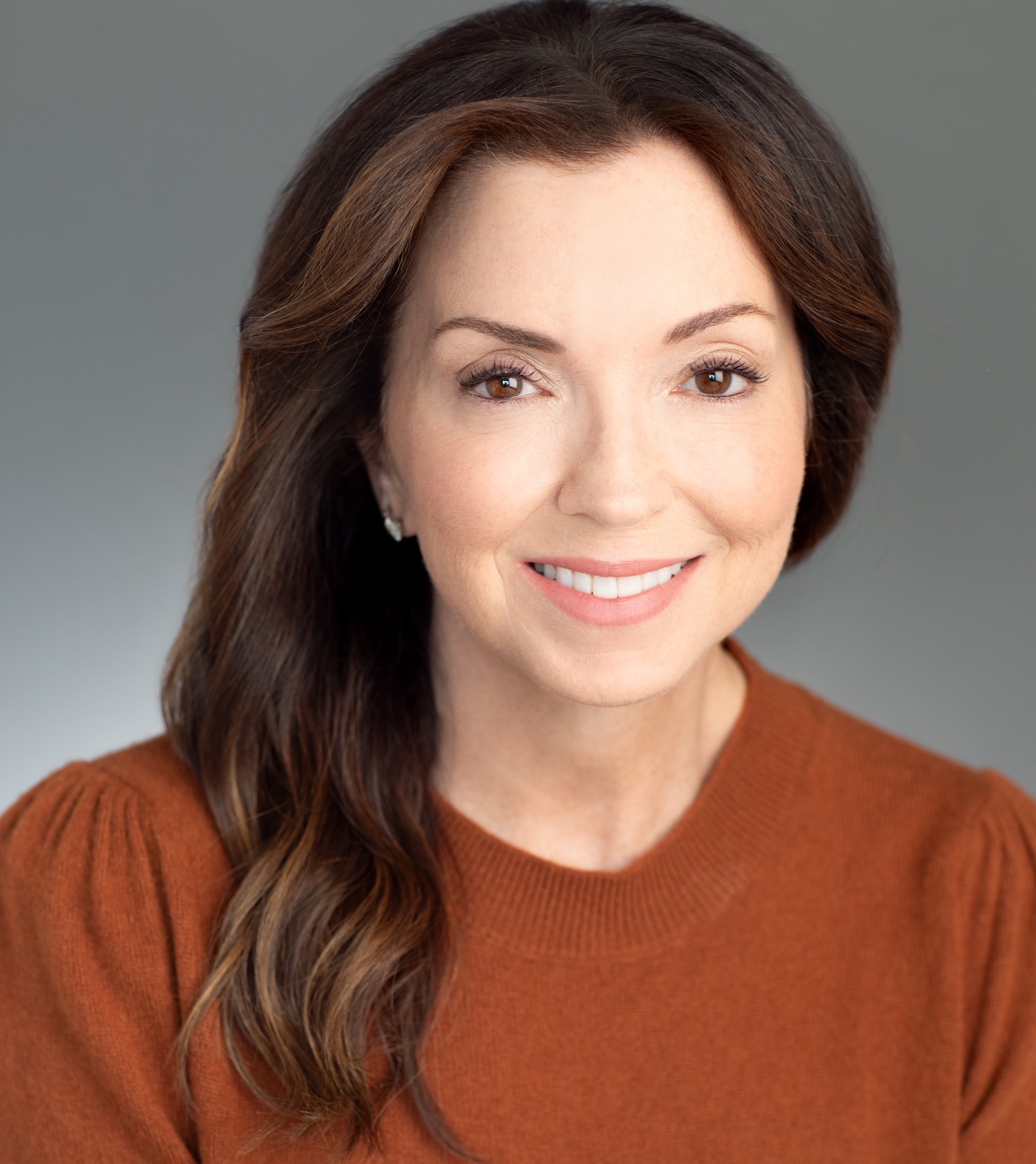 Woman with long brown hair wearing a rust-colored top, smiling at the camera against a plain gray background.