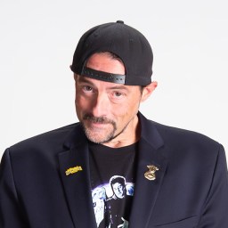 Man wearing a black backward baseball cap, dark blazer with pins, and a graphic T-shirt, standing against a plain white background.