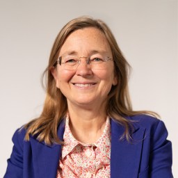 A woman with long light brown hair, glasses, and a blue blazer over a patterned shirt, smiling at the camera against a plain light background.