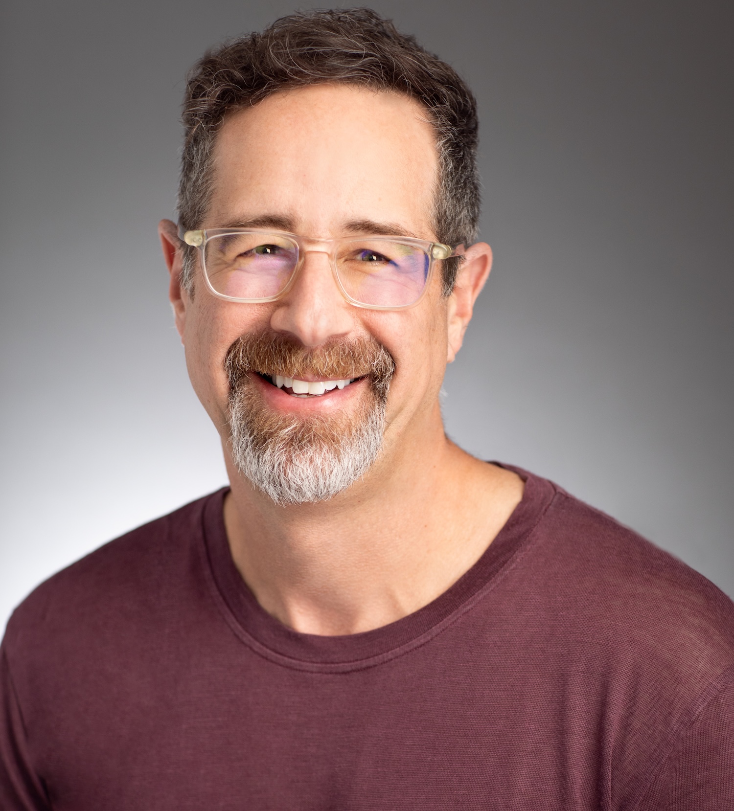 A middle-aged man with glasses, short curly hair, and a salt-and-pepper beard, wearing a maroon shirt, smiling in front of a gray background.