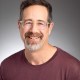 A middle-aged man with glasses, short curly hair, and a salt-and-pepper beard, wearing a maroon shirt, smiling in front of a gray background.