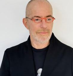 A bald man with a short grey beard wears red glasses, a black jacket, and a black shirt, standing against a plain white background.