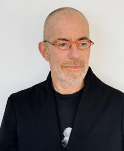 A bald man with a short grey beard wears red glasses, a black jacket, and a black shirt, standing against a plain white background.