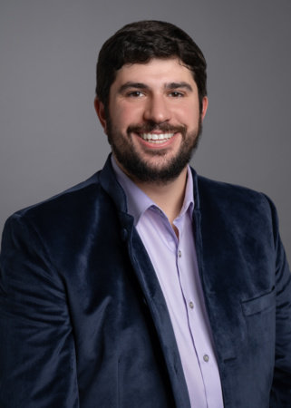 Man with short dark hair and beard, wearing a blue velvet jacket over a light purple shirt, smiles in front of a plain gray background.