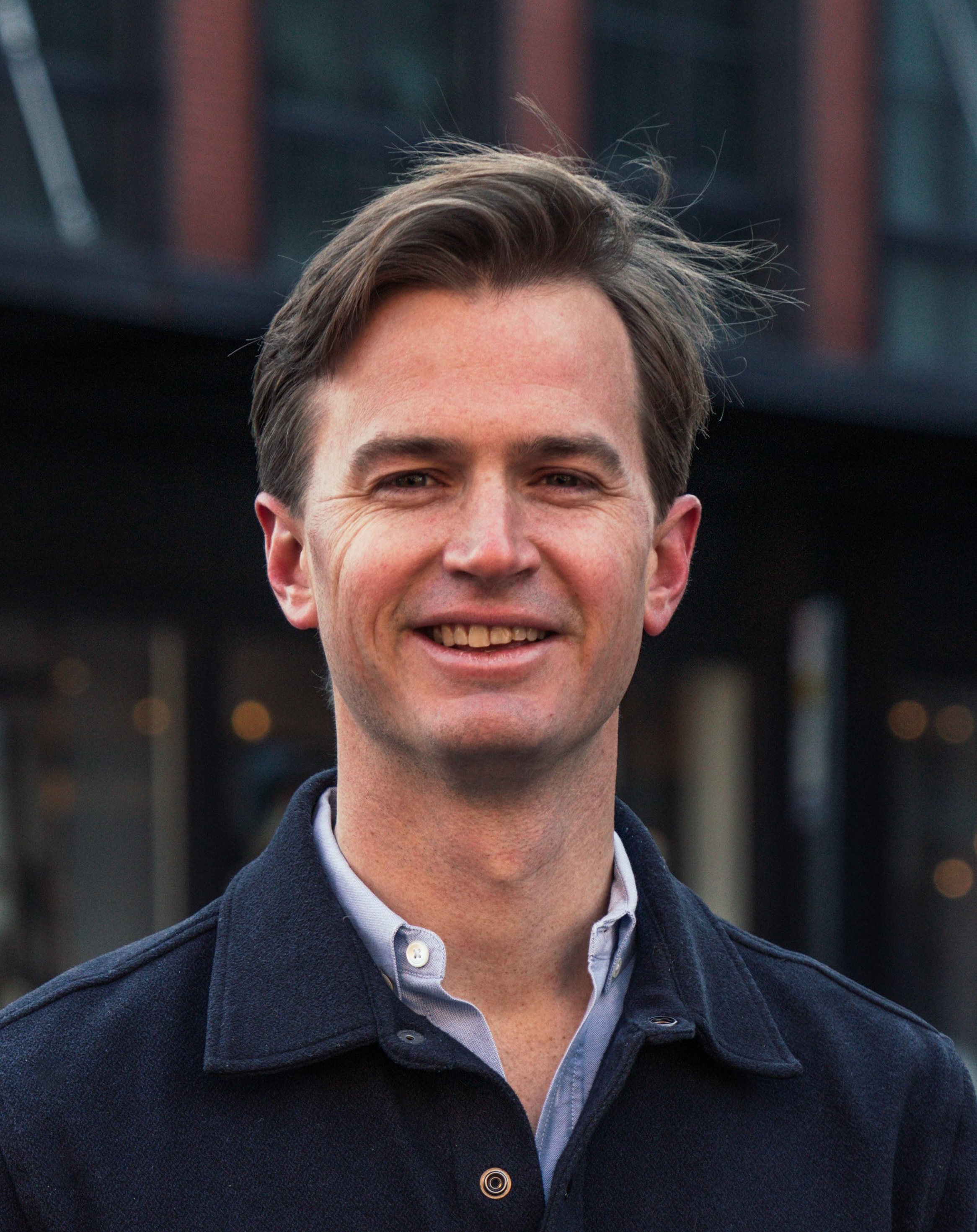 A man with light brown hair, wearing a dark jacket over a light shirt, stands outdoors and smiles at the camera.